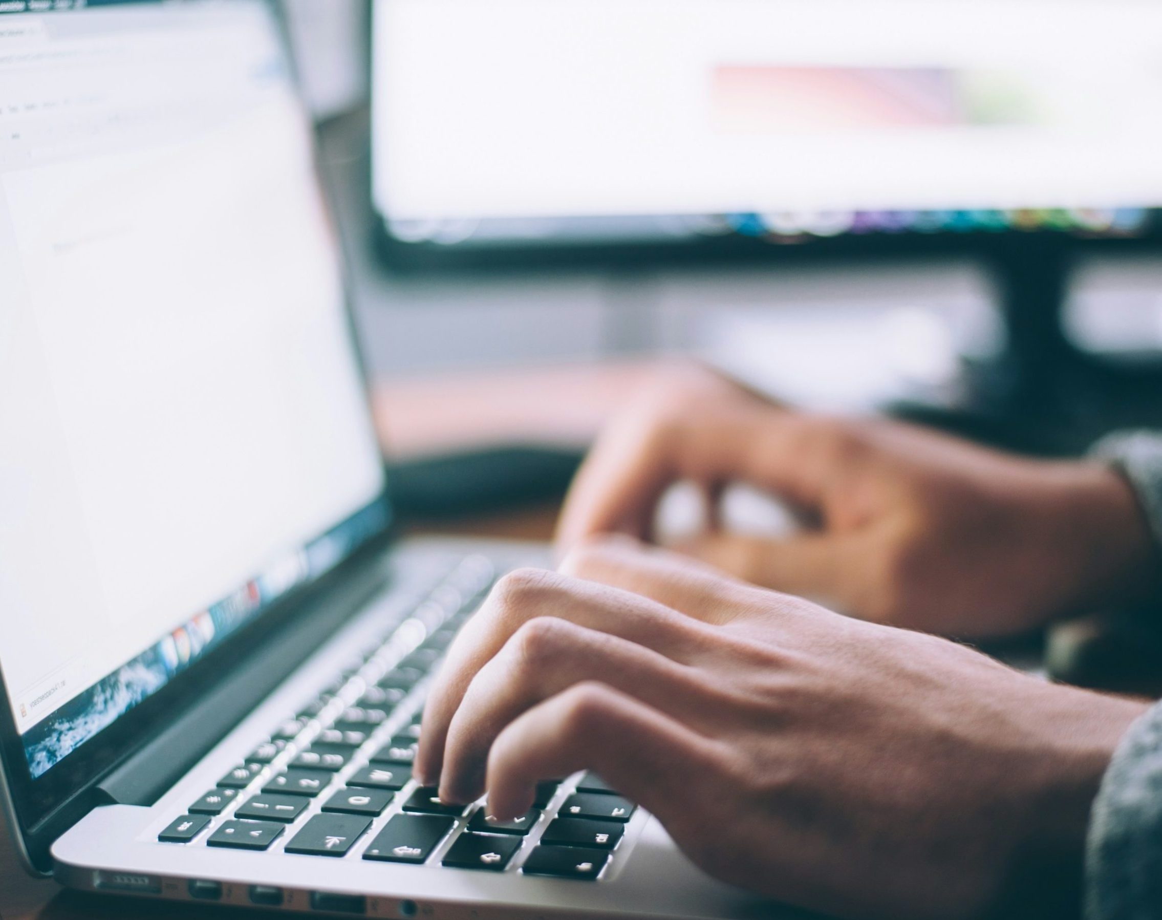 Hands typing on a laptop with another screen in the background